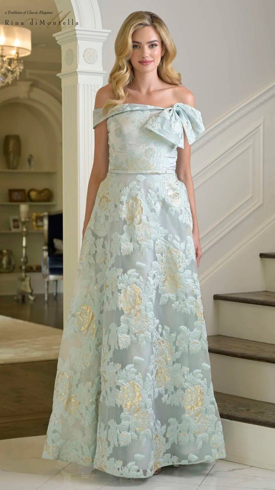 A woman stands indoors on a staircase, wearing the Rina Di Montella RD3401 long formal dress in Seaglass, featuring an off-shoulder design with a pale blue A-line skirt, gold and white floral embroidery, and a large bow on the shoulder.